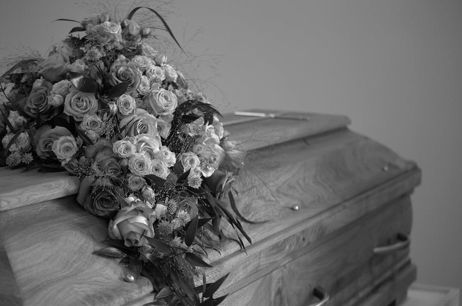 A funeral flower arrangement placed atop a closed wooden casket, featuring a large bouquet of fresh roses in various shades, including light and dark tones, complemented by greenery and delicate fille