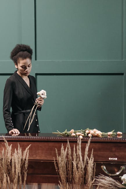 A woman with dark skin and curly hair, dressed in a black blazer, is holding a single white flower with a pink center and green stem. She stands next to a wooden casket draped with a floral arrangemen
