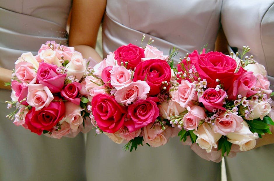 A floral arrangement consisting of three bouquets of fresh roses held by a person wearing a light-colored dress, with roses in shades of pink, blush, and deep red. The bouquets feature tightly cluster