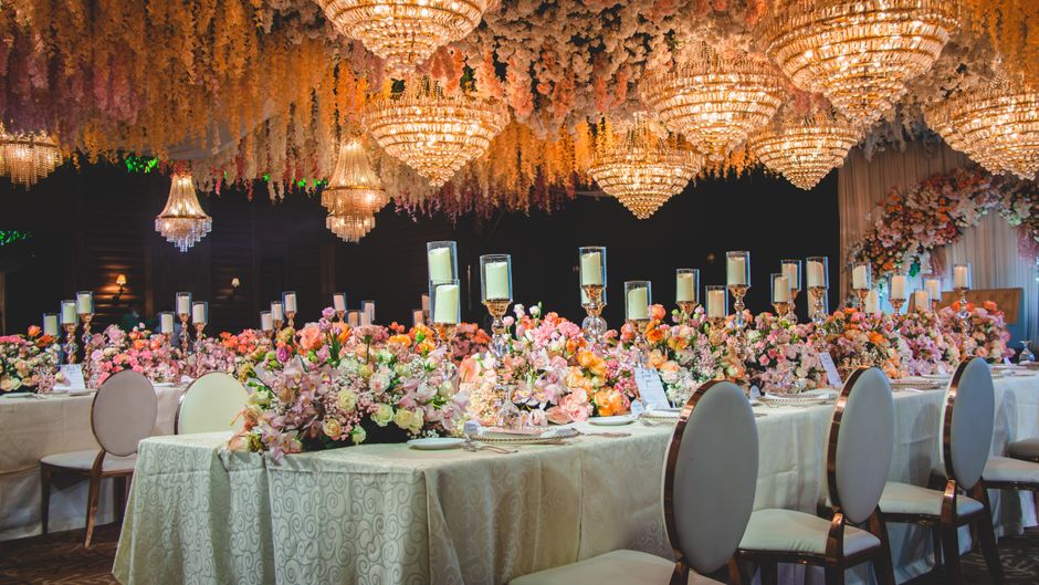 An elegant banquet hall decorated with a long, cream-colored table arrangement featuring a variety of fresh flowers in soft pastel shades including pinks, peach, cream, and white. The floral centerpie