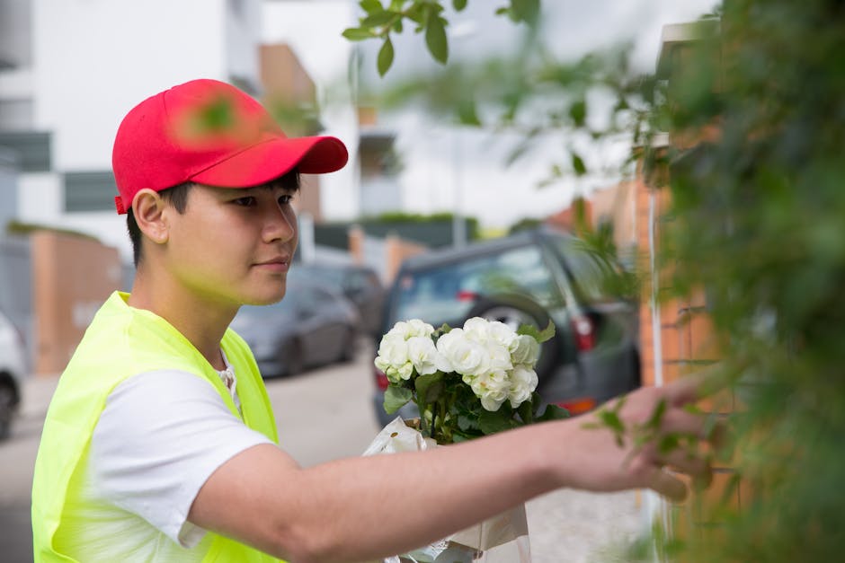 A young male florist wearing a red cap and a high-visibility yellow vest is holding a bouquet of white roses and hydrangeas wrapped in white paper, preparing a floral gift outdoors on a residential st
