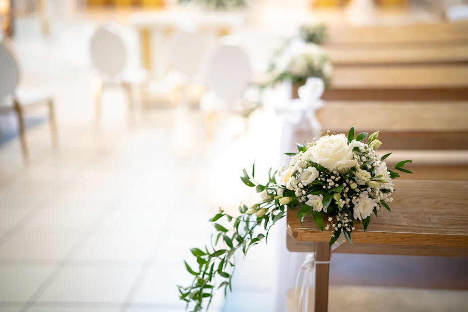 A beautifully arranged bridal bouquet featuring white roses, baby's breath, and lush green foliage, resting on a wooden pew in a softly lit wedding venue. The bouquet is wrapped with elegant white rib