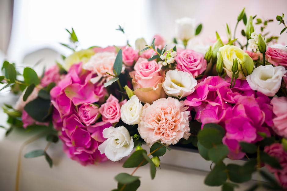 A floral arrangement featuring a lush bouquet of fresh flowers, including pink and white roses, cream-colored eustoma, and soft pink carnations, all interwoven with dark green eucalyptus leaves. The b